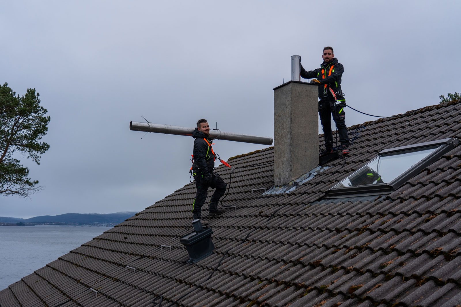 To personer på taket som jobber med pipemontering. En står ved pipa, og den andre bærer på et rør.